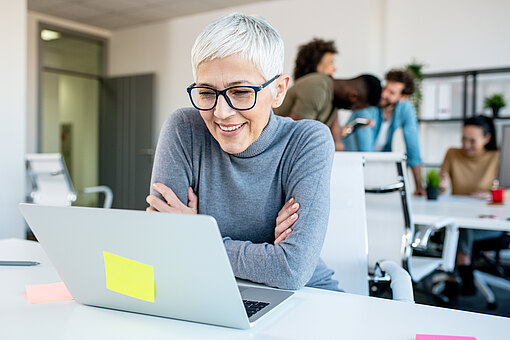 Eine ältere Frau mit kurzen grauen Haaren und Brille lächelt, während sie an einem Laptop in einem Büro arbeitet.