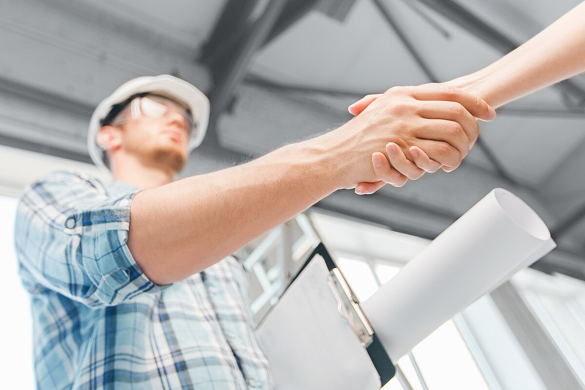 Ein Bauarbeiter mit Helm und Bauplänen schüttelt einer anderen Person die Hand auf einer Baustelle