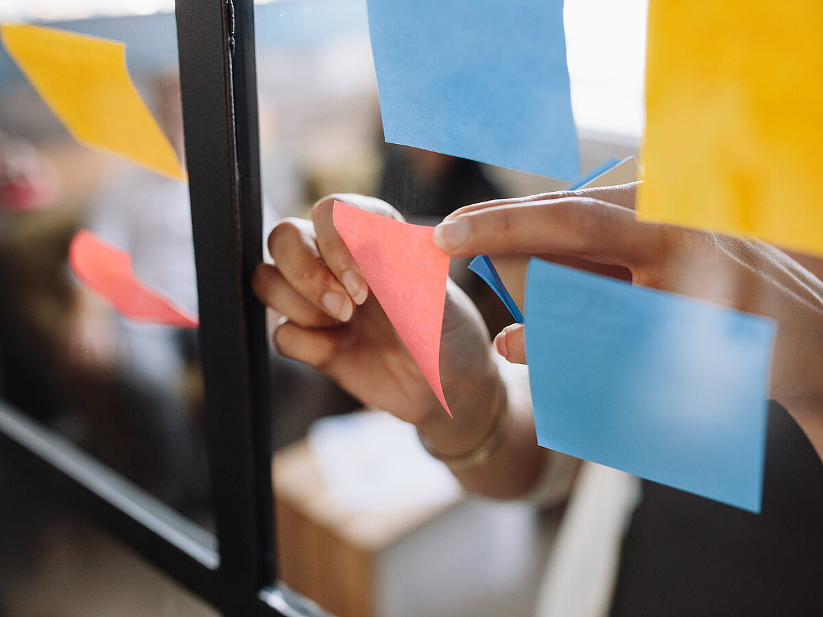 Hände befestigen bunte Haftnotizen an einer Glaswand während eines Meetings