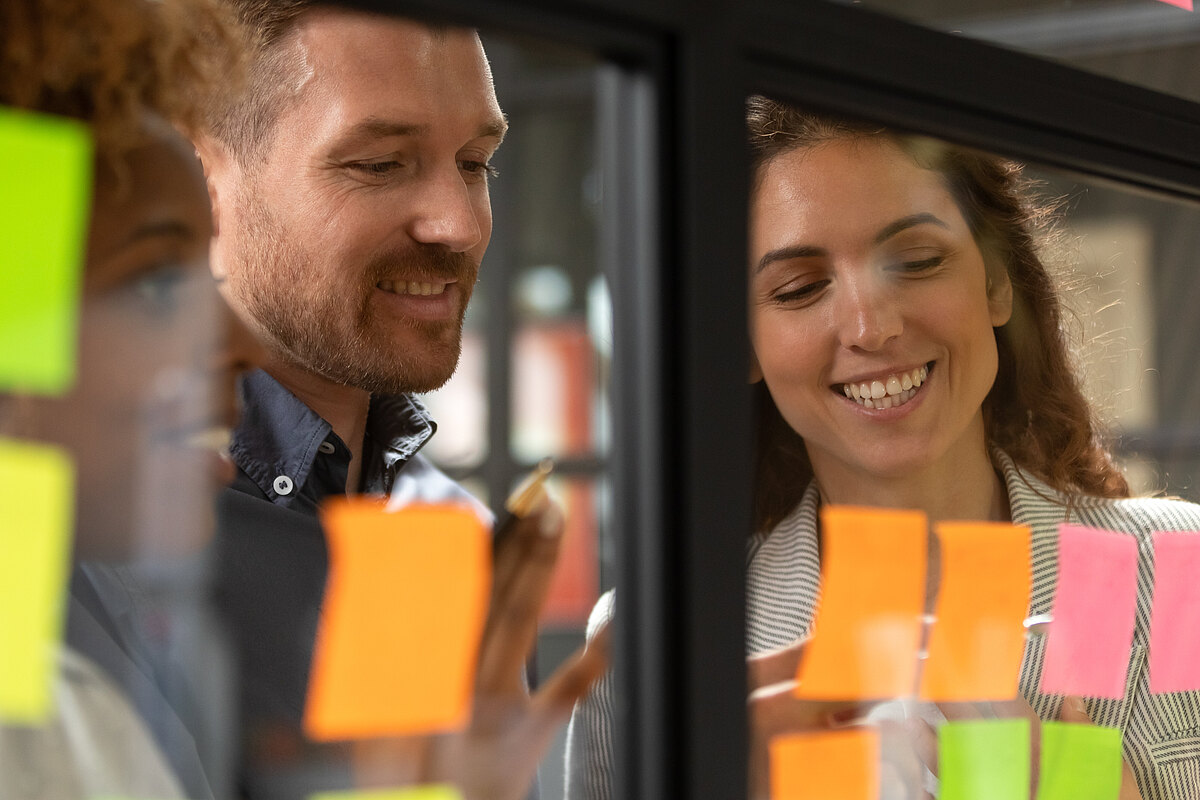 Drei lächelnde Büroangestellte arbeiten an einem Projekt mit Haftnotizen an einer Glaswand