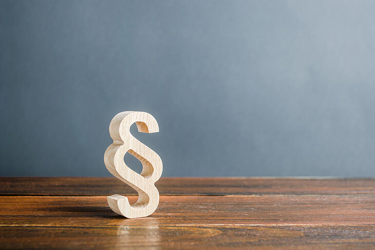 Ein hölzernes Paragraphenzeichen steht auf einem Holztisch vor einer grauen Wand