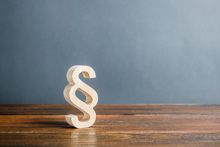 Ein hölzernes Paragraphenzeichen steht auf einem Holztisch vor einer grauen Wand