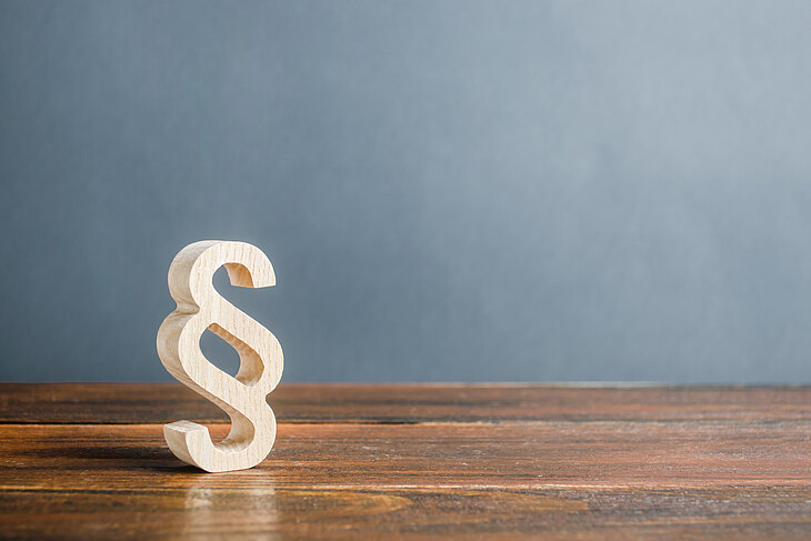 Ein hölzernes Paragraphenzeichen steht auf einem Holztisch vor einer grauen Wand