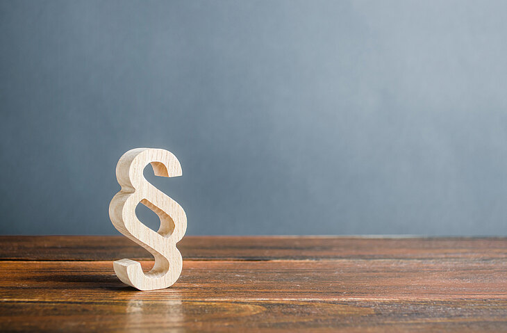 Ein hölzernes Paragraphenzeichen steht auf einem Holztisch vor einer grauen Wand