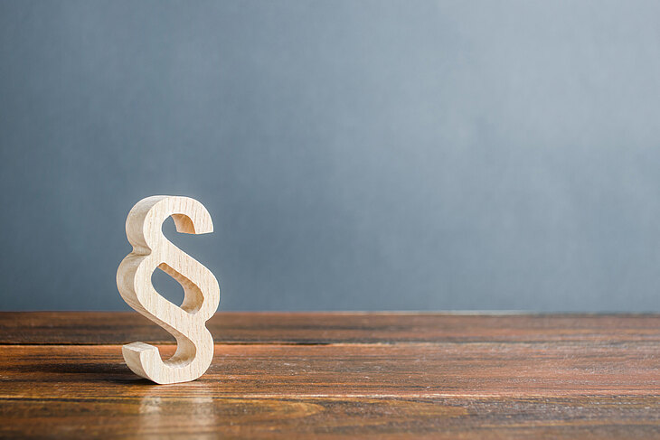 Ein hölzernes Paragraphenzeichen steht auf einem Holztisch vor einer grauen Wand