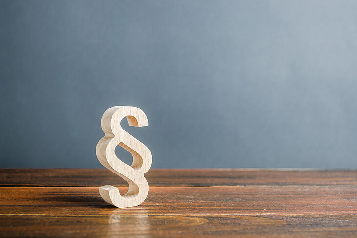 Ein hölzernes Paragraphenzeichen steht auf einem Holztisch vor einer grauen Wand