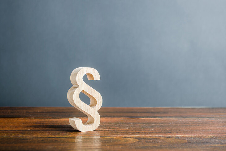 Ein hölzernes Paragraphenzeichen steht auf einem Holztisch vor einer grauen Wand