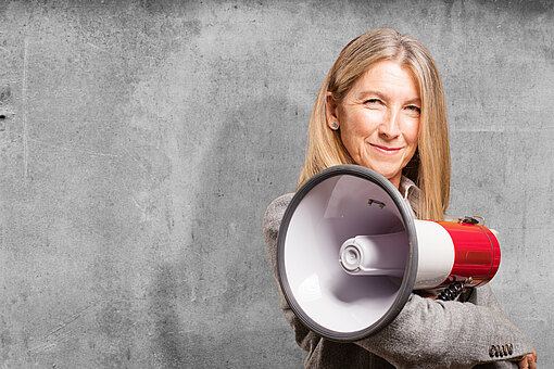 Selbstbewusste Frau mit langen blonden Haaren hält ein rotes Megafon vor grauer Betonwand