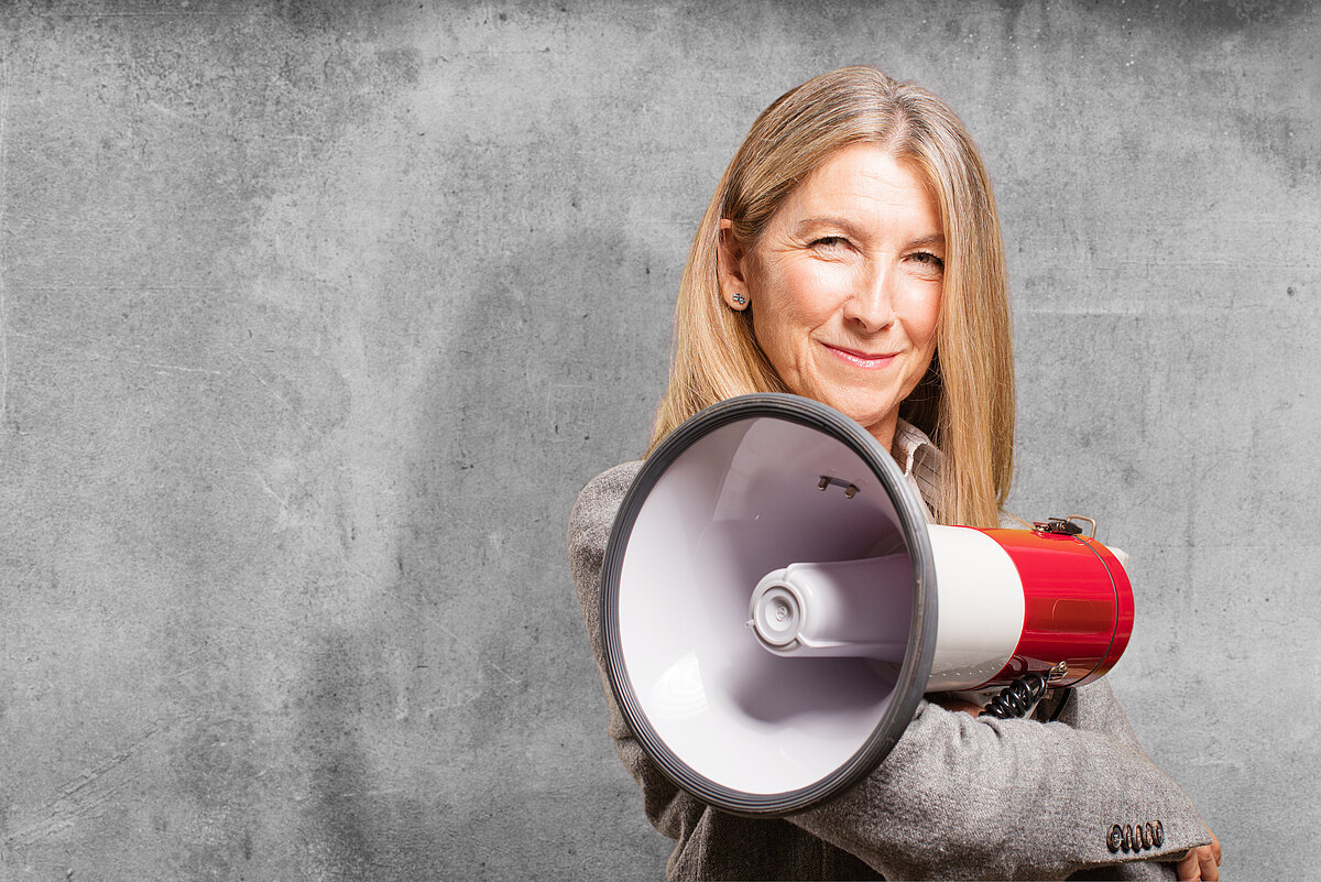 Selbstbewusste Frau mit langen blonden Haaren hält ein rotes Megafon vor grauer Betonwand